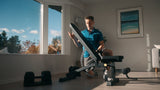 Man exercising on a home fitness machine in a room with large windows and a couch.
